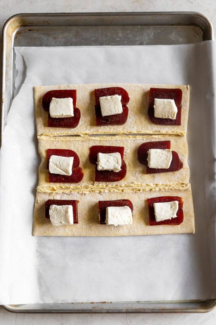 Nine squares of puff pastry dough on a parchment-lined baking sheet, prepared like pastelitos de guayaba, each topped with a slice of quince paste and a cube of white cheese.