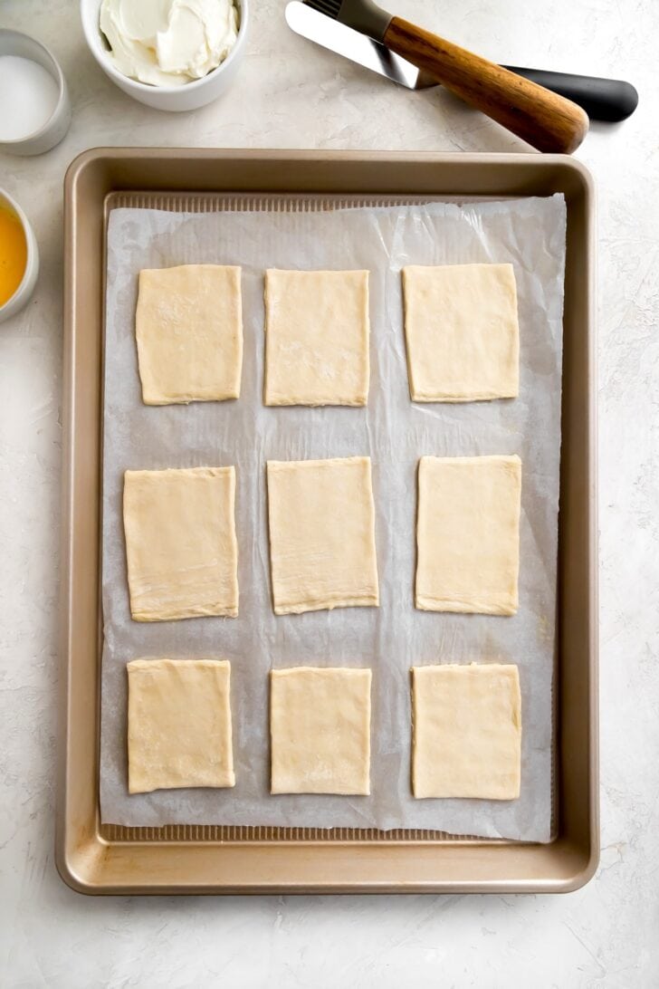 Nine rectangular pieces of raw puff pastry for pastelitos de queso arranged in three rows on a parchment-lined baking sheet.