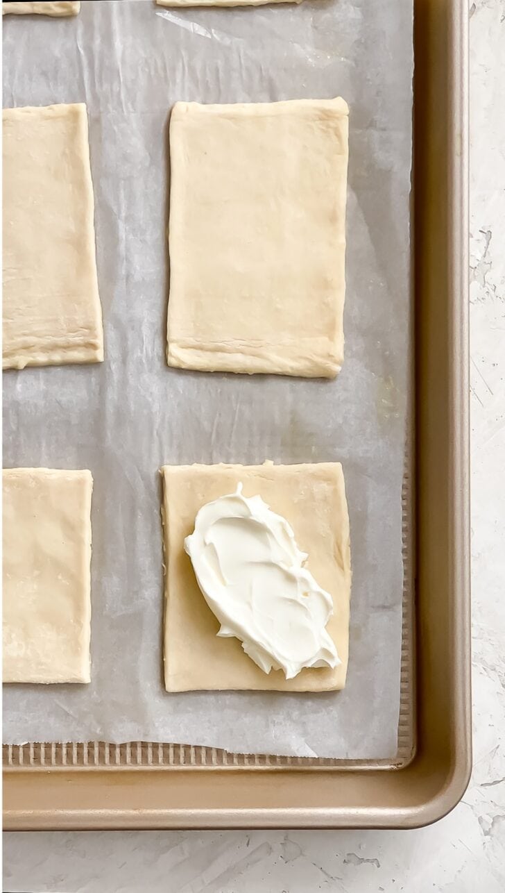 Rectangular pieces of raw puff pastry arranged on a parchment-lined baking sheet to make pastelitos de queso, with a dollop of cream cheese spread onto one of the pieces.