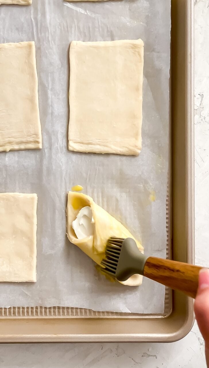 A hand uses a silicone brush to apply egg wash to folded pastelitos de queso filled with cream cheese on a parchment-lined baking sheet.
