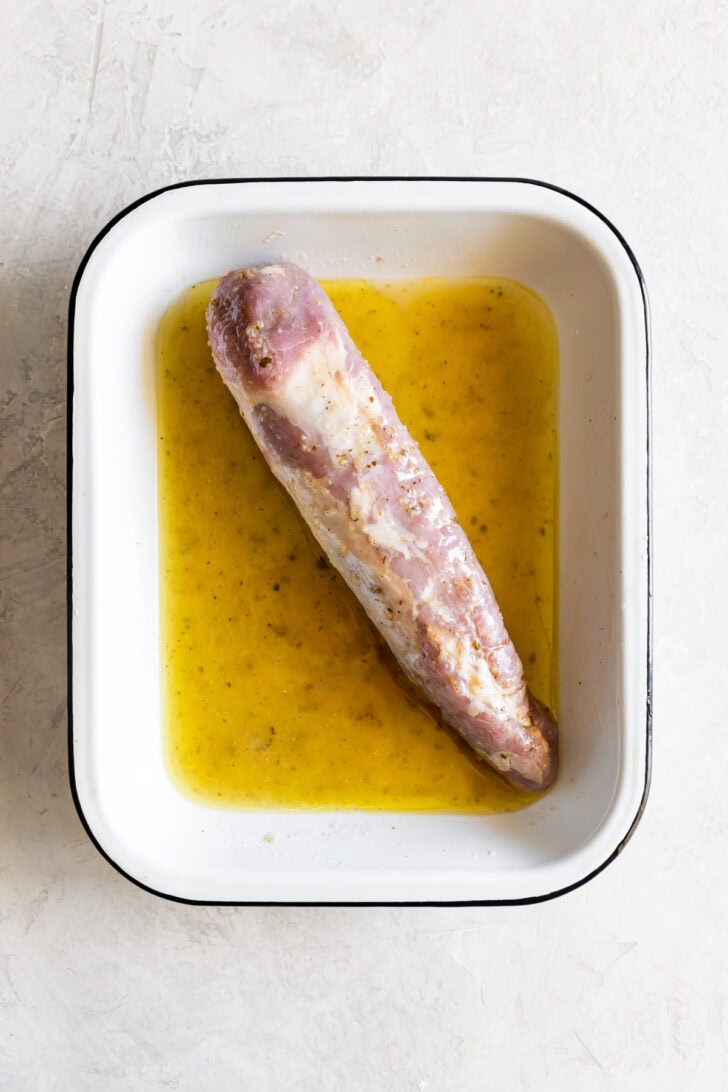 A raw pork tenderloin for a Cuban sandwich marinating in a yellow liquid inside a white rectangular baking dish.