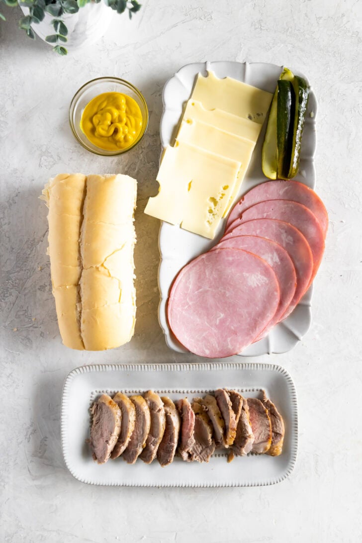 Ingredients for a Cuban sandwich, including a loaf of bread, sliced ham, roasted pork, Swiss cheese, a pickle spear, and a bowl of mustard.