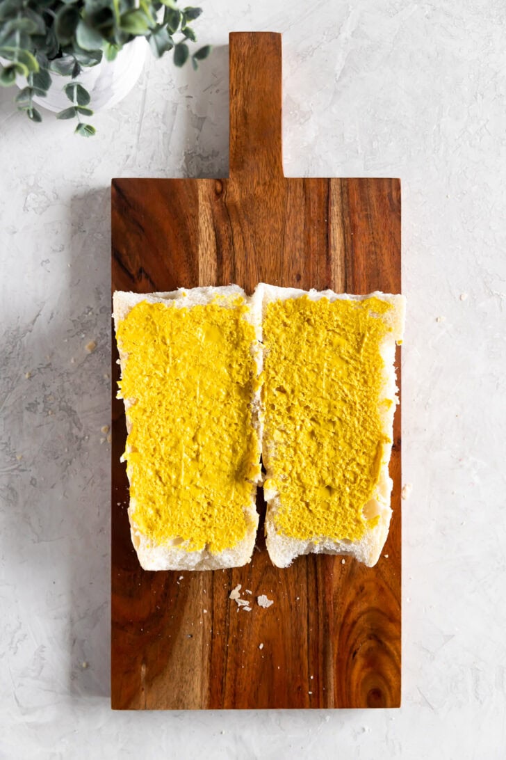 Two halves of a Cuban sandwich roll spread with yellow mustard on a wooden cutting board.