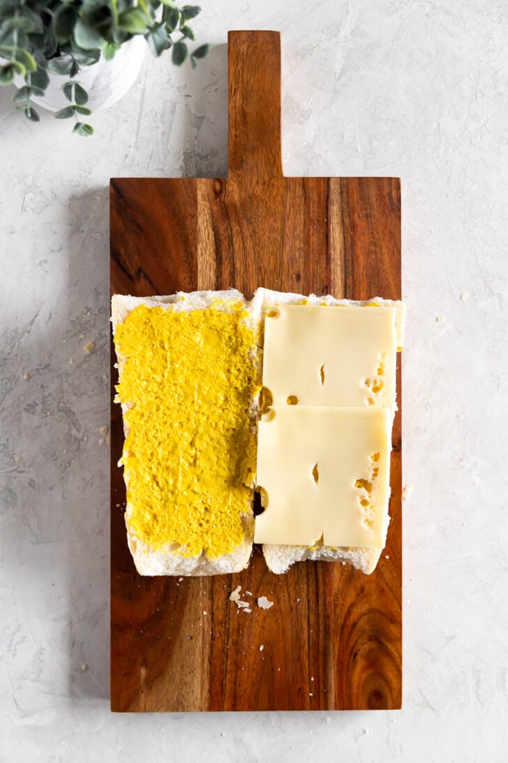 Two open halves of a Cuban sandwich roll on a wooden board, one spread with yellow mustard and the other topped with two slices of Swiss cheese.