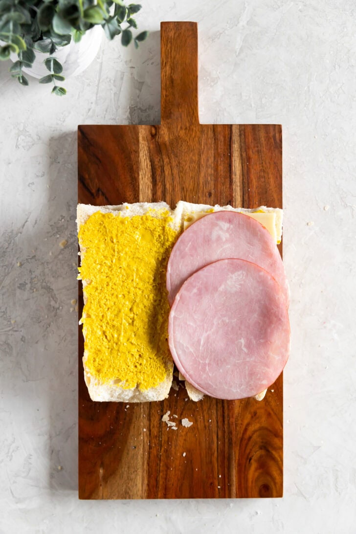 Open-faced Cuban sandwich on a wooden board with one half spread with yellow mustard and the other topped with ham and cheese.
