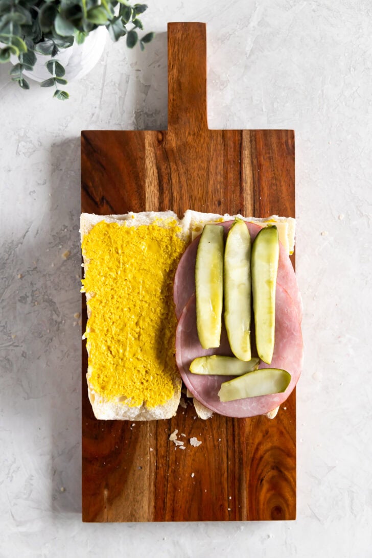 A Cuban sandwich on a wooden board, featuring one piece of bread spread with yellow mustard and another topped with ham, cheese, and pickle slices.