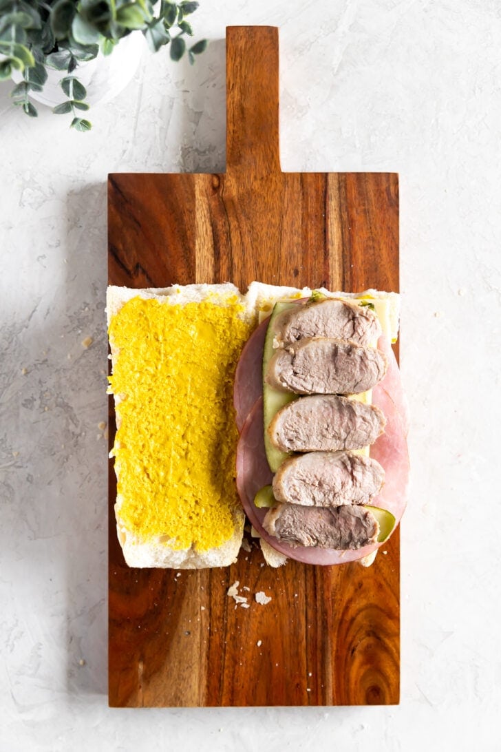 An open-faced Cuban sandwich on a wooden board, featuring a mustard-spread baguette on one side and ham, sliced pork, and pickles on the other.