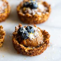 Granola cups made with oats, almonds and walnuts, filled with cashew butter caramel then topped with blueberries, shredded coconut, and lemon zest.