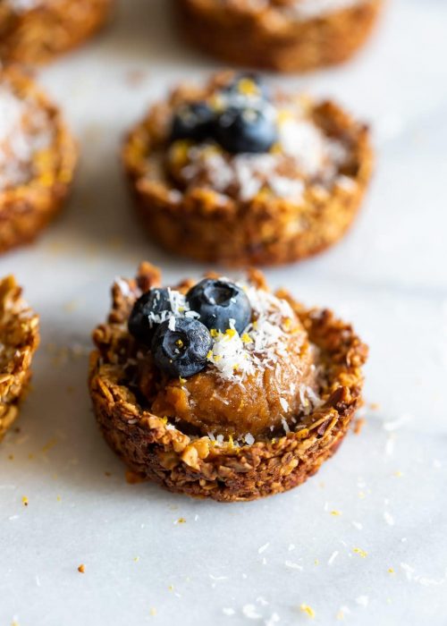 Granola cups made with oats, almonds and walnuts, filled with cashew butter caramel then topped with blueberries, shredded coconut, and lemon zest.