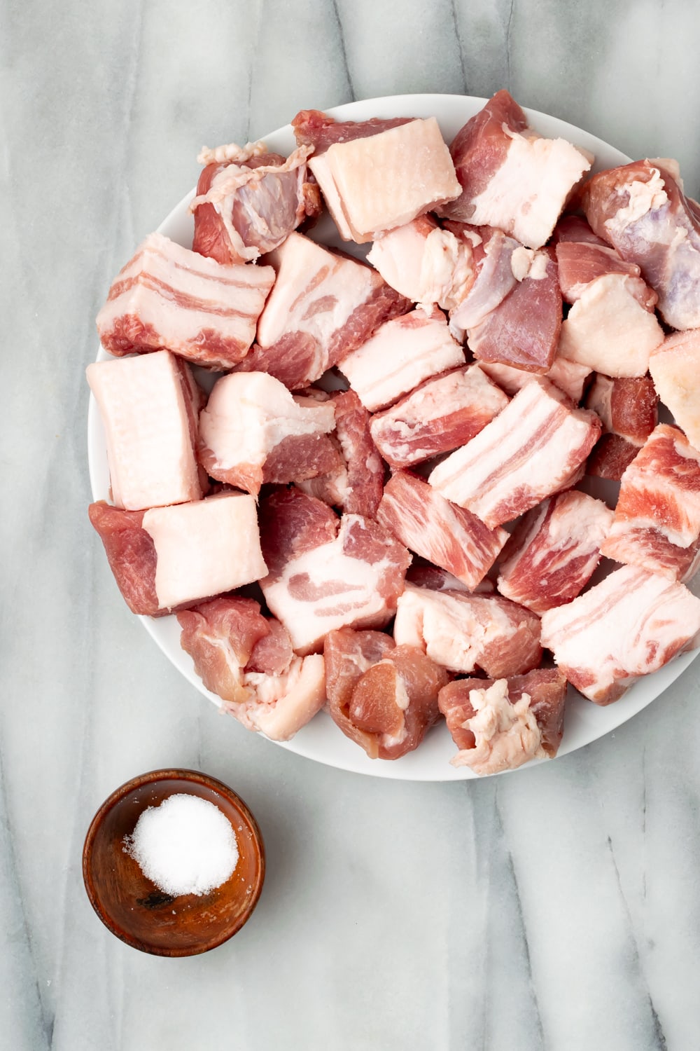 Cut pork belly pieces in 1 inch cubes on a round white plate with a small wooden bowl of salt on the side.