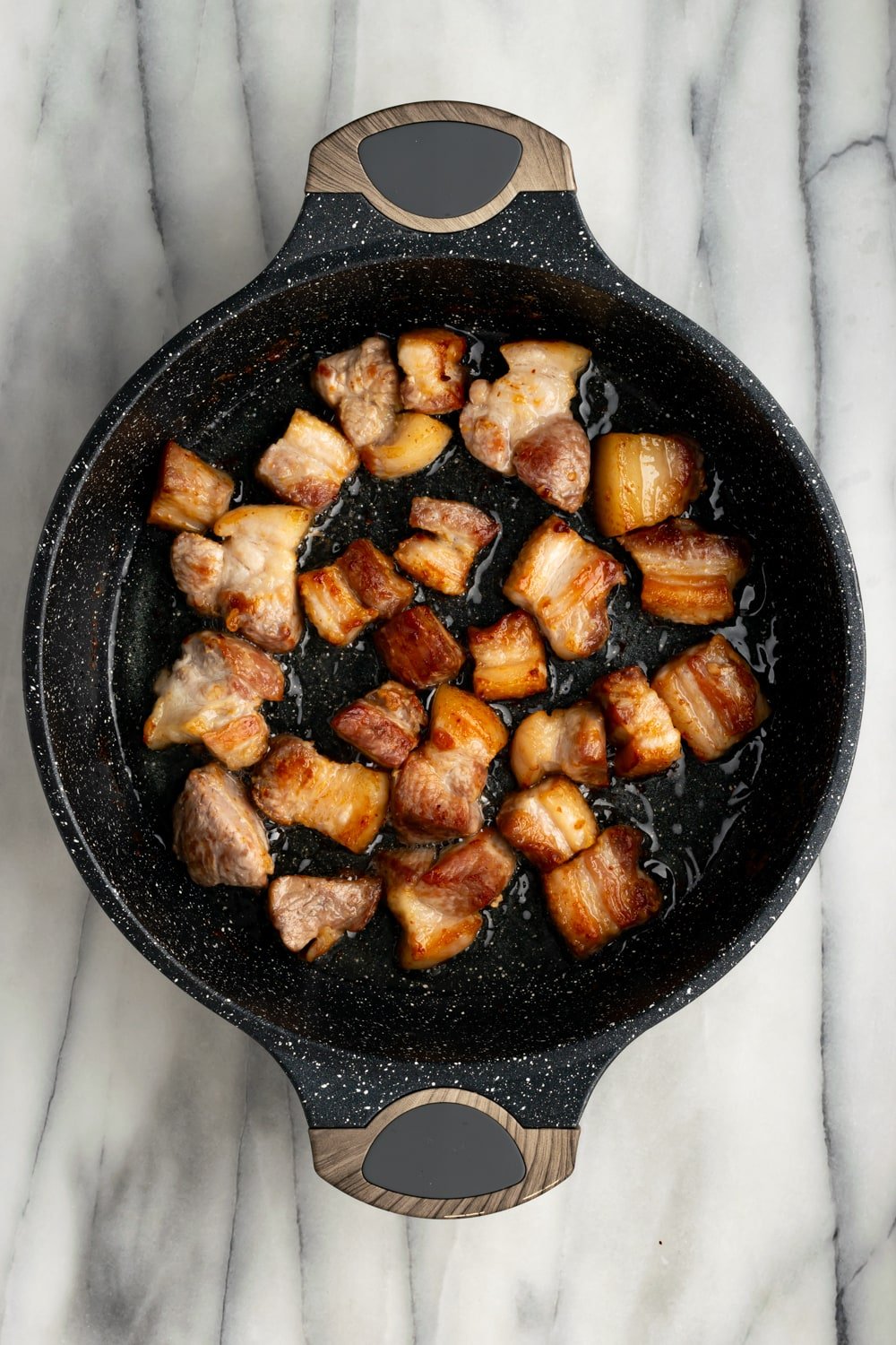 Pork belly pieces cooking in a cast iron pan to make chicharrones de puerco on a marble countertop