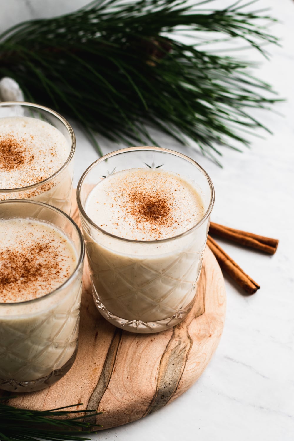 coquito drink recipe in glasses with a sprinkle of cinnamon on a wood board on a white surface with cinnamon sticks