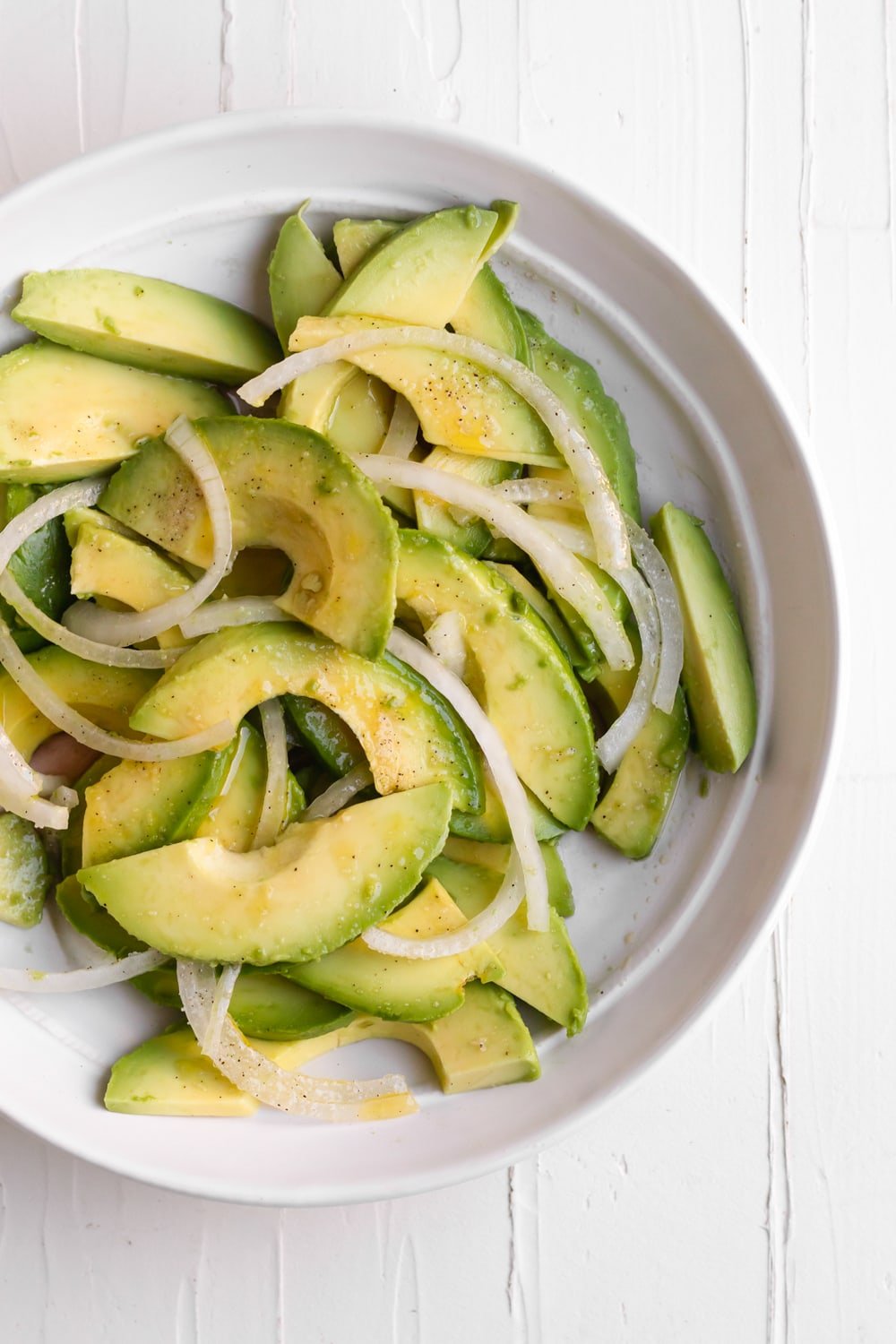 a white plate with sliced avocado, sliced onions, oil and vinegar, salt and pepper