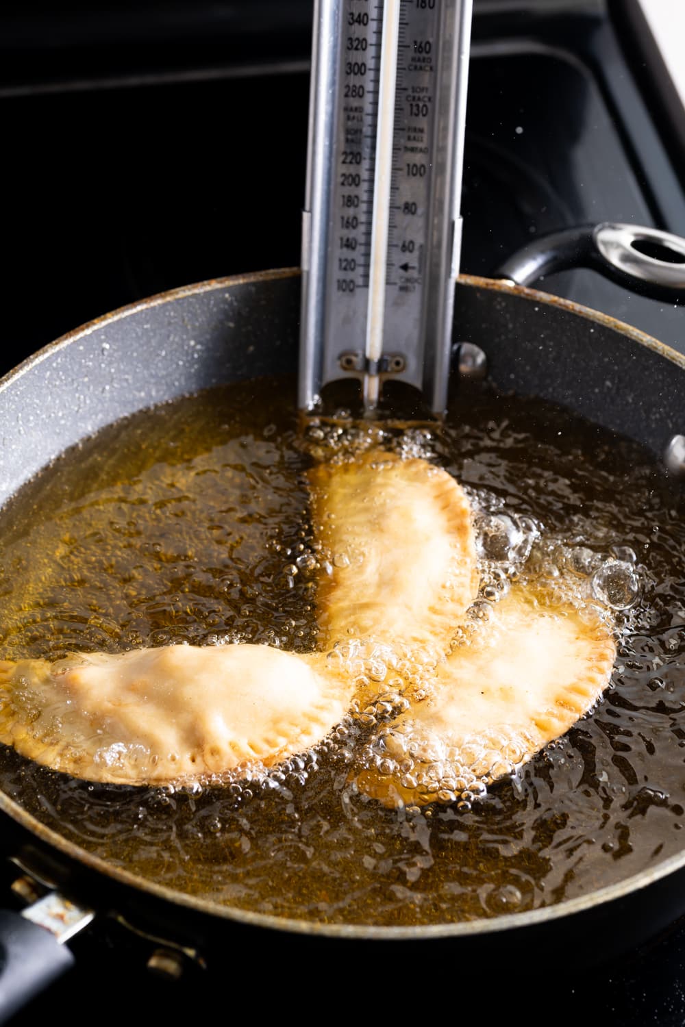 Three cuban chicken empanadas being fried in oil in a deep fry pan with a thermometer in the oil
