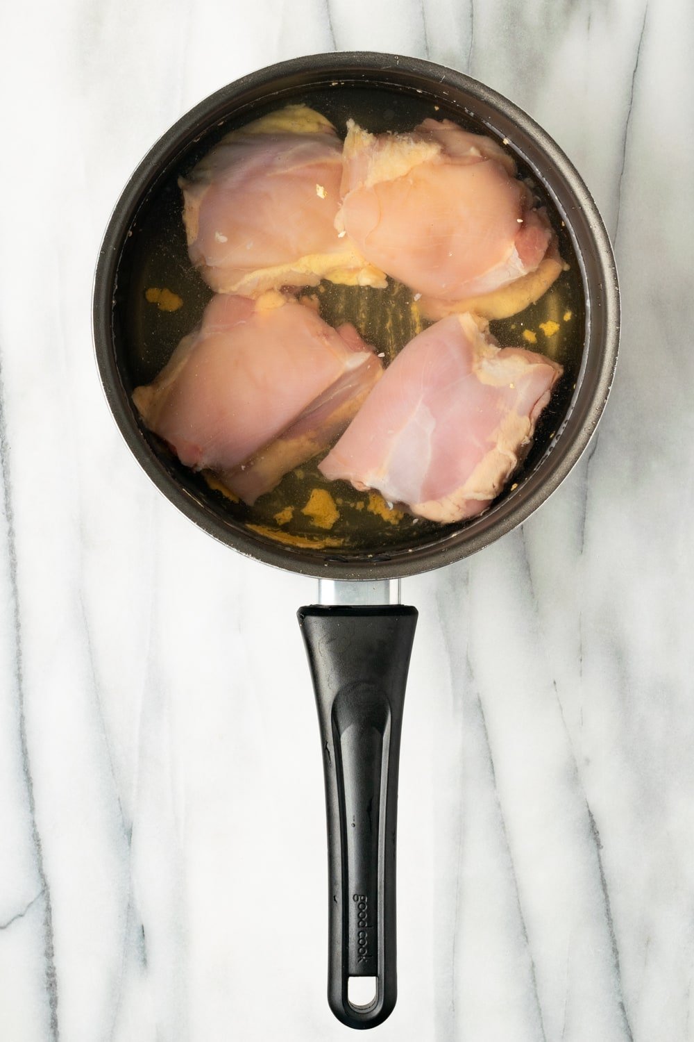 An overhead shot of four chicken thighs in a saucepan in water with chicken bouillon in it on a marble background