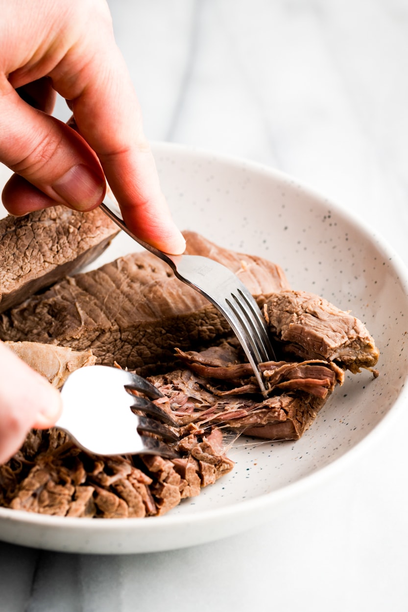 boiled flank steak being shredded with two forks on a white plate.