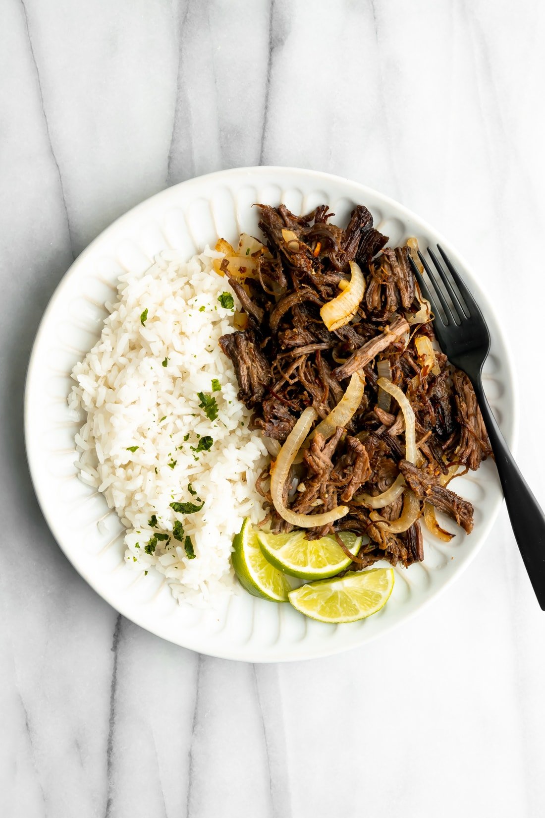 shredded fried beef with white rice and lime wedges on a white plate on a marble surface
