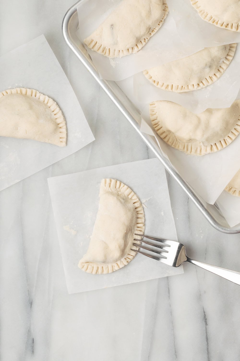 A guava cheese empanada being sealed with a fork with more uncooked empanadas on a baking sheet on the side.