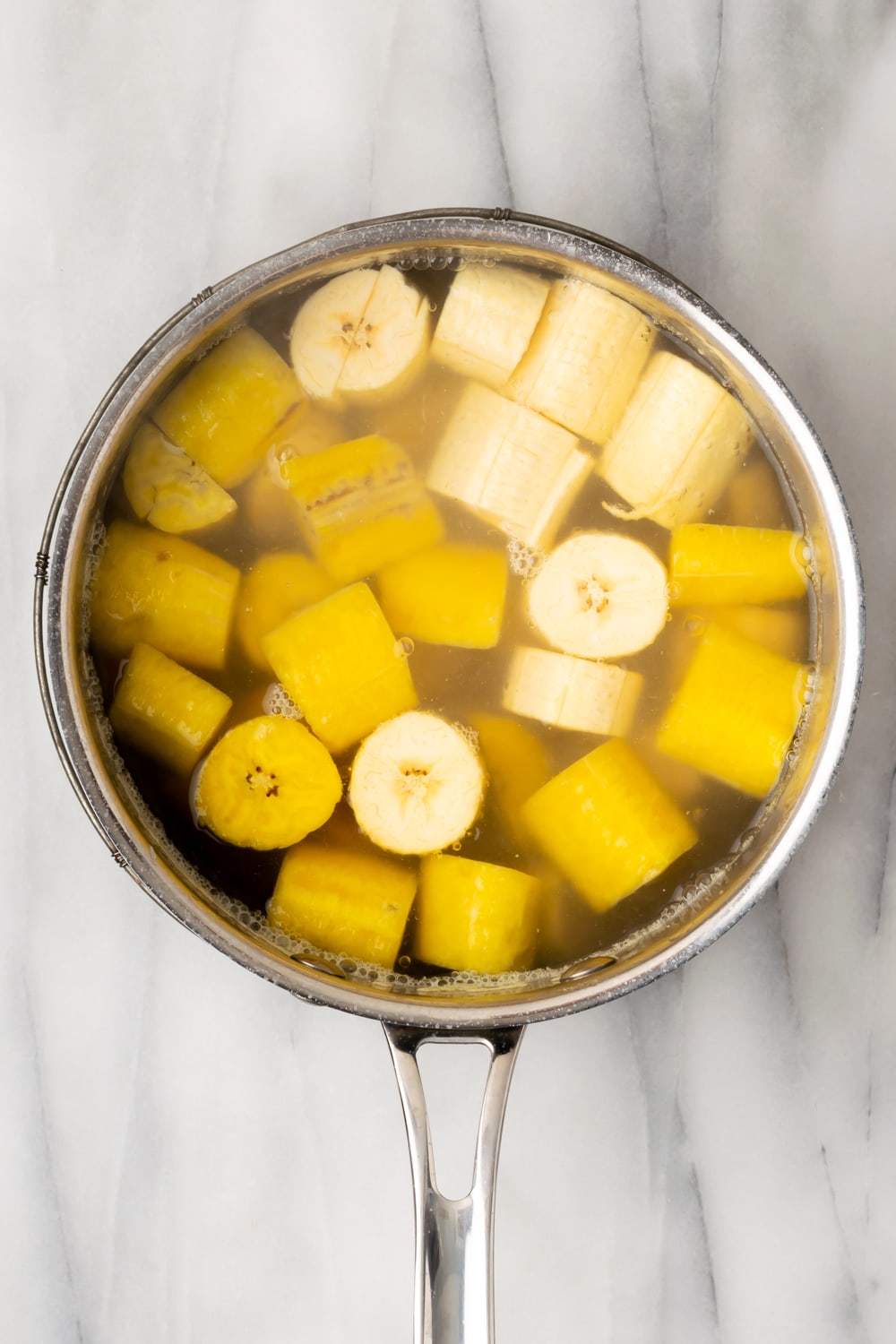 Sliced yellow and green plantains in a saucepan filled with water on a marble background. 