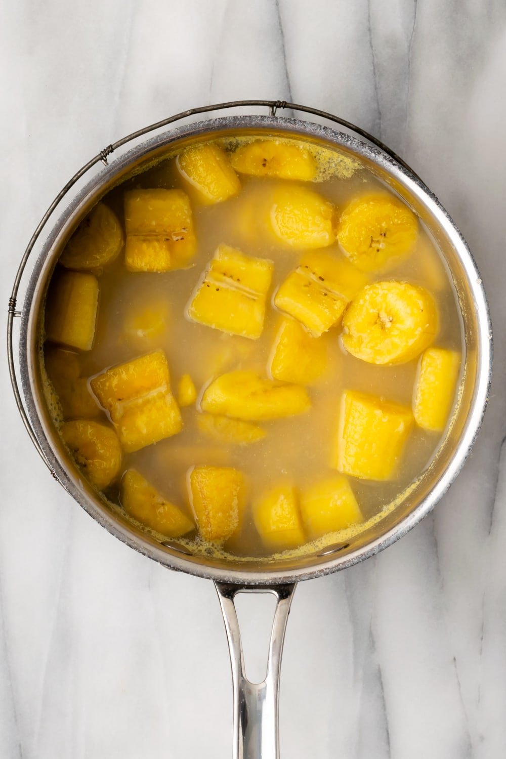 Boiled/softened sliced yellow and green plantains in a saucepan filled with water on a marble background.