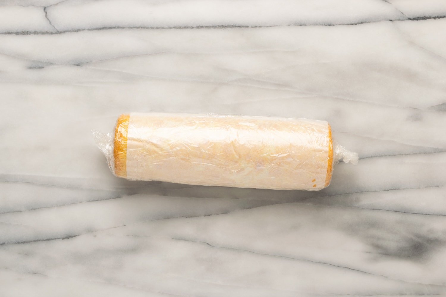 A guava swiss roll that has been rolled up and wrapped tightly in parchment paper and plastic wrap on a marble counter