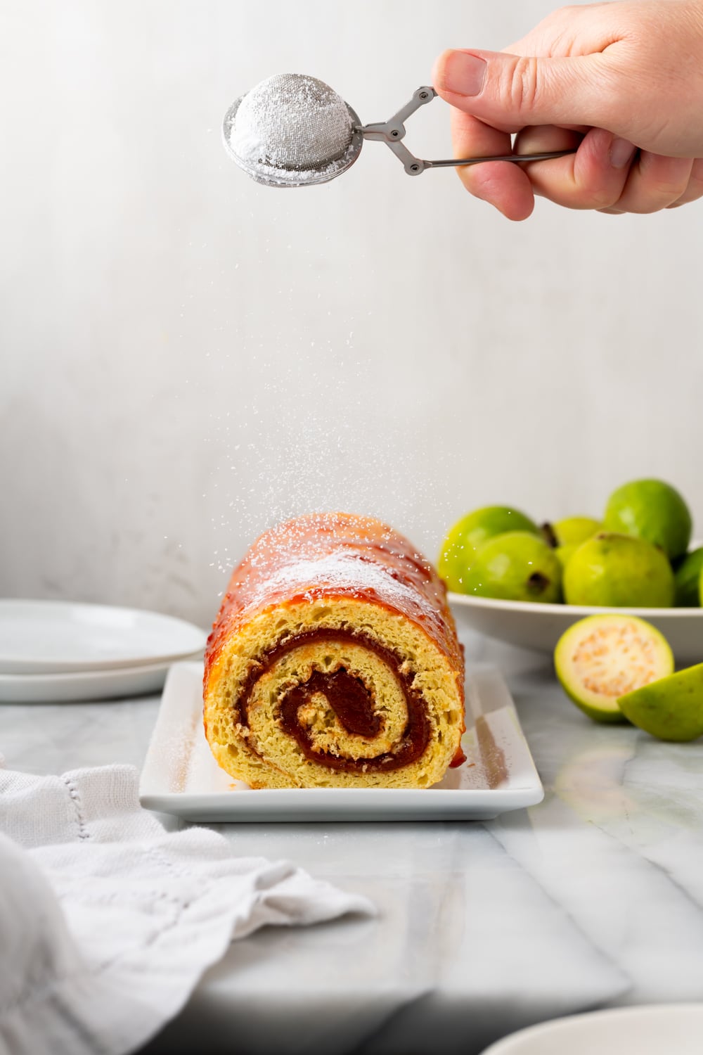 A guava swiss roll on a white platter that is being dusted with powdered sugar with a bowl of whole guavas in the background