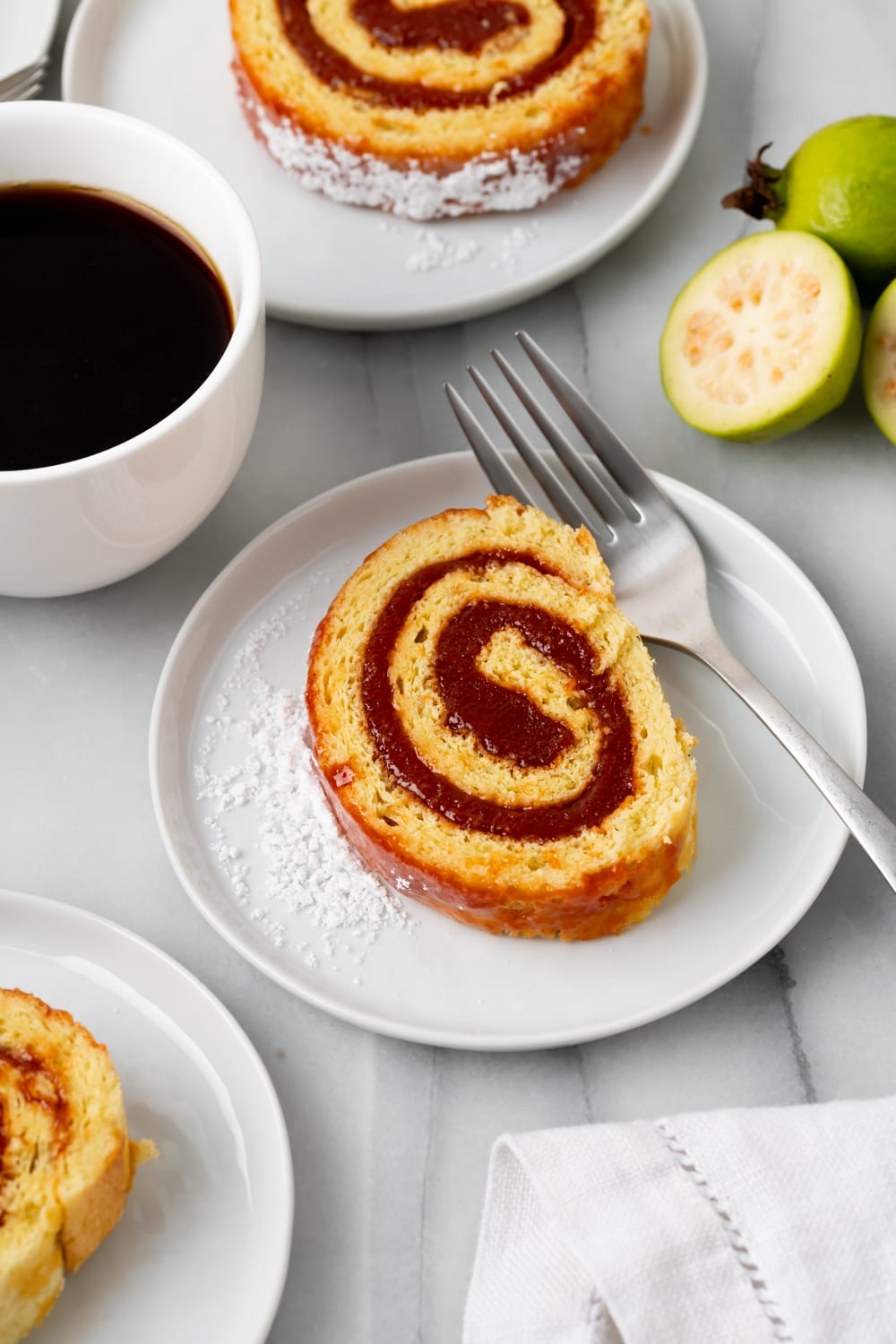 A piece of a guava swiss roll cake on a white plate with a fork on it with a white mug of coffee on the side
