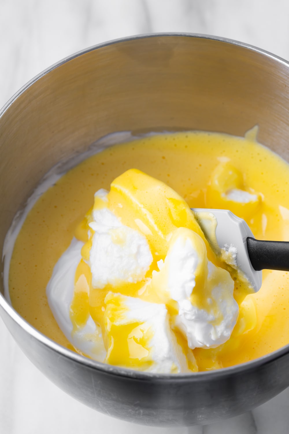 Egg white mixture being folded into a beat egg yolk mixture with a spatula all in a silver bowl to make the batter for brazo gitano