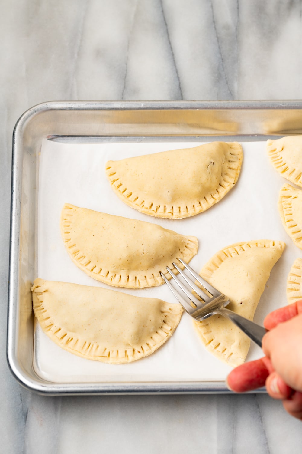 A fork showing how to make empanada dough fully sealed with other empanadas on the same parchment lined baking sheet