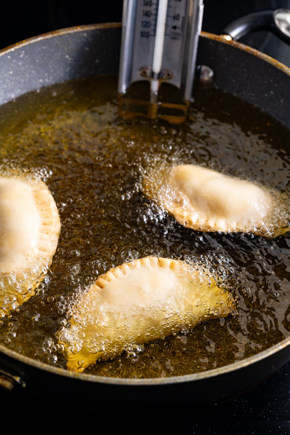 Three beef filled cuban empanadas being fried in oil in a deep fry pan with a thermometer in the oil
